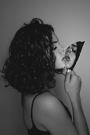 A black and white portrait features a person with curly hair holding a broken mirror shard. The reflection in the shard shows part of their face, creating a dramatic and introspective effect.
