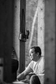 A man is sitting on a porch, looking contemplative. He is wearing a t-shirt and appears to be in a relaxed, thoughtful mood. Behind him, a wooden panel structure with natural light filtering through makes the setting appear serene. A small bag or camera case hangs from one of the beams.