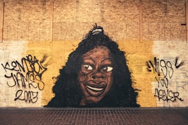 A mural painted on a wooden surface depicts a large, stylized portrait of a person with curly hair and a wide smile. Graffiti text surrounds the portrait, with messages such as 'Miss You 4Ever' and a name. The colors are earthy, with a background of brown tones and black outlines.
