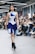 A fashion model walks down a runway wearing a unique asymmetrical dress with bold blue and white geometric patterns. The setting is an indoor fashion show with an audience seated on either side, some of whom are taking pictures.