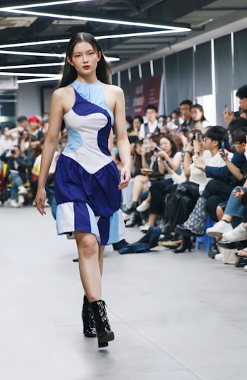 A fashion model walks down a runway wearing a unique asymmetrical dress with bold blue and white geometric patterns. The setting is an indoor fashion show with an audience seated on either side, some of whom are taking pictures.