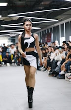 A person confidently walks down a runway in a crowded room, wearing a stylish black and white outfit with high platform boots. The audience, seated on either side of the runway, appears engaged, with some individuals capturing the moment on their phones. Overhead lighting adds a modern and sleek atmosphere to the setting.