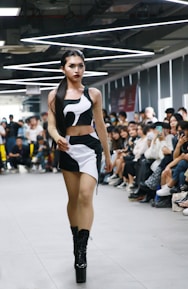 A person confidently walks down a runway in a crowded room, wearing a stylish black and white outfit with high platform boots. The audience, seated on either side of the runway, appears engaged, with some individuals capturing the moment on their phones. Overhead lighting adds a modern and sleek atmosphere to the setting.