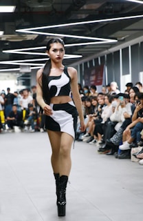 A person confidently walks down a runway in a crowded room, wearing a stylish black and white outfit with high platform boots. The audience, seated on either side of the runway, appears engaged, with some individuals capturing the moment on their phones. Overhead lighting adds a modern and sleek atmosphere to the setting.
