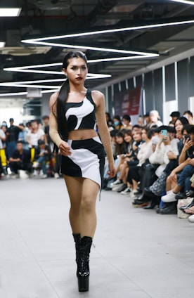 A person confidently walks down a runway in a crowded room, wearing a stylish black and white outfit with high platform boots. The audience, seated on either side of the runway, appears engaged, with some individuals capturing the moment on their phones. Overhead lighting adds a modern and sleek atmosphere to the setting.