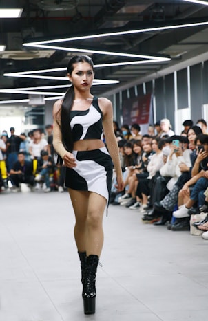 A person confidently walks down a runway in a crowded room, wearing a stylish black and white outfit with high platform boots. The audience, seated on either side of the runway, appears engaged, with some individuals capturing the moment on their phones. Overhead lighting adds a modern and sleek atmosphere to the setting.