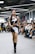 A person confidently walks down a runway in a crowded room, wearing a stylish black and white outfit with high platform boots. The audience, seated on either side of the runway, appears engaged, with some individuals capturing the moment on their phones. Overhead lighting adds a modern and sleek atmosphere to the setting.