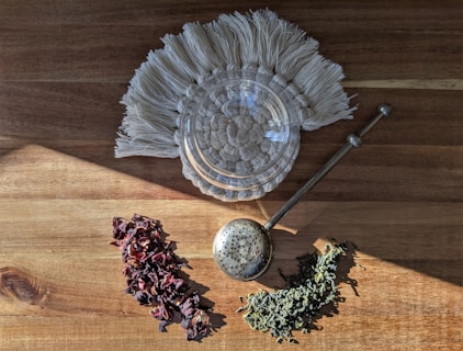 On a wooden surface, there are sprigs of dried herbs and an infusion spoon. A braided circular mat holds a glass jar, which sits near a decorative item with fringes.