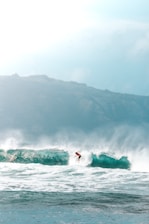 A surfer skillfully rides a large ocean wave, creating an impressive display of balance and agility. The backdrop features a rugged coastline under a bright, slightly overcast sky.