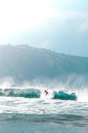 A surfer skillfully rides a large ocean wave, creating an impressive display of balance and agility. The backdrop features a rugged coastline under a bright, slightly overcast sky.