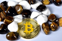 A gold-colored Bitcoin coin is surrounded by polished stones. The stones are a mix of white and tiger's eye gemstones, which have a rich brown and yellow hue.