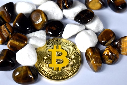 A gold-colored Bitcoin coin is surrounded by polished stones. The stones are a mix of white and tiger's eye gemstones, which have a rich brown and yellow hue.