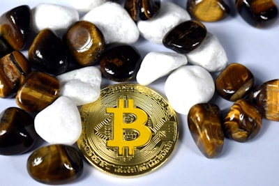 A gold-colored Bitcoin coin is surrounded by polished stones. The stones are a mix of white and tiger's eye gemstones, which have a rich brown and yellow hue.
