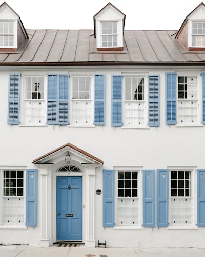 A two-story white house with a metal roof and three dormer windows. The facade features numerous windows with decorative blue shutters. The central entrance has a blue door with white trim and a small overhang supported by columns.