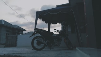 A motorcycle is parked under a canopy in a residential area with overcast skies in the background. The setting appears to be a concrete patio or driveway, with buildings visible on either side.
