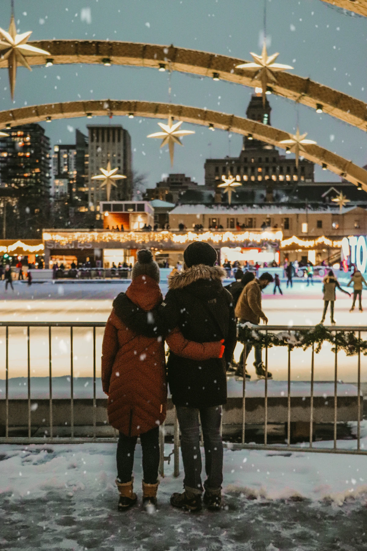 Couple standing at the rail of an outdoor ice rink with lit market stalls behind them