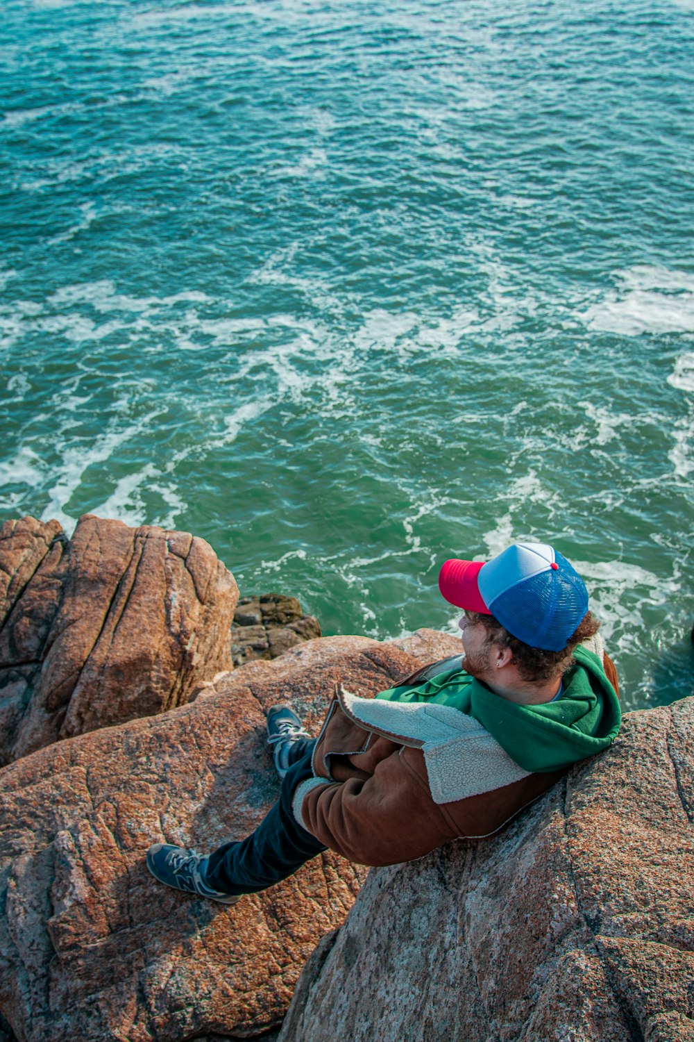 man in brown jacket and blue cap sitting on brown rock formation near body of water