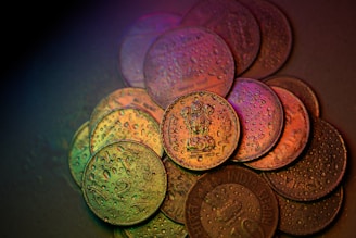 A close-up view of several Indian coins arranged on a surface, covered in water droplets. The coins display various denominations and details, with a multicolored lighting effect creating a gradient of hues over their surfaces.