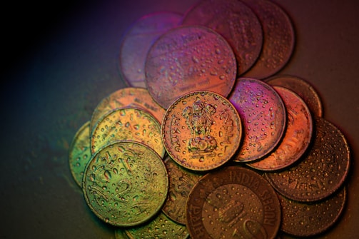 A close-up view of several Indian coins arranged on a surface, covered in water droplets. The coins display various denominations and details, with a multicolored lighting effect creating a gradient of hues over their surfaces.