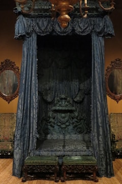 An ornate, vintage canopy bed adorned with intricate blue drapery and detailed wood carvings. The drapes are heavy and richly patterned, framing the bed and creating a sense of enclosure. The bed is accompanied by two small upholstered stools with wooden legs featuring elaborate carvings. Two oval mirrors with decorative frames flank the bed on the wall, and the setting is warm, with a chandelier overhead adding a touch of grandeur.