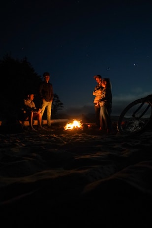 A group of riders gathered around their bikes under a starry sky, sharing stories by a campfire.