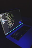 A laptop displays a programming code editor on its screen, slightly tilted with a dark background. The screen glows with various colored code lines, predominantly green, blue, and white. The keyboard is visible, casting a blue hue, suggesting a low-light environment.