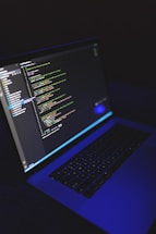 A laptop displays a programming code editor on its screen, slightly tilted with a dark background. The screen glows with various colored code lines, predominantly green, blue, and white. The keyboard is visible, casting a blue hue, suggesting a low-light environment.