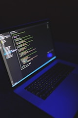 A laptop displays a programming code editor on its screen, slightly tilted with a dark background. The screen glows with various colored code lines, predominantly green, blue, and white. The keyboard is visible, casting a blue hue, suggesting a low-light environment.