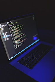 A laptop displays a programming code editor on its screen, slightly tilted with a dark background. The screen glows with various colored code lines, predominantly green, blue, and white. The keyboard is visible, casting a blue hue, suggesting a low-light environment.