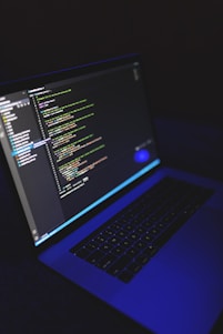 A laptop displays a programming code editor on its screen, slightly tilted with a dark background. The screen glows with various colored code lines, predominantly green, blue, and white. The keyboard is visible, casting a blue hue, suggesting a low-light environment.