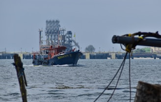 A blue and red boat with the name 'Muharam' is sailing on the water. In the background, there is an industrial structure with pipes and metal frameworks, and beyond that, a large bridge. The scene has a calm atmosphere with minimal waves on the water surface.