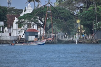 A traditional fishing net setup extends over the water near a vibrant fishing boat. The backdrop features a tree-lined shoreline with colonial-style architecture, including a white building with spires and a red-roofed shed. Several people are visible along the shoreline.