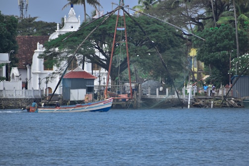 A traditional fishing net setup extends over the water near a vibrant fishing boat. The backdrop features a tree-lined shoreline with colonial-style architecture, including a white building with spires and a red-roofed shed. Several people are visible along the shoreline.