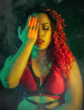 A person with curly hair colored in vibrant red and black wearing a red top with black straps, covering one eye with their hand. The lighting is dramatic with neon green and red tones, and there is a smoky or misty ambiance.