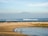 A serene beach scene with two children playing near the water's edge. In the background, gentle waves roll in, and a large mountain rises under a blue sky. A few boats can be seen on the horizon while the sandy shore blends into rocky outcrops.