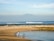 A serene beach scene with two children playing near the water's edge. In the background, gentle waves roll in, and a large mountain rises under a blue sky. A few boats can be seen on the horizon while the sandy shore blends into rocky outcrops.