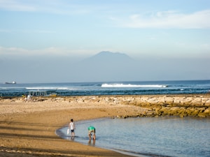 A serene beach scene with two children playing near the water's edge. In the background, gentle waves roll in, and a large mountain rises under a blue sky. A few boats can be seen on the horizon while the sandy shore blends into rocky outcrops.