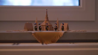 A small, detailed wooden figurine scene featuring miniature houses and people, likely depicting a historical or village setting. The figures are dressed in traditional attire with blue hats and black clothing. The structures include two small houses and one larger building with a steeple, flanked by ornamental sheaves of wheat.