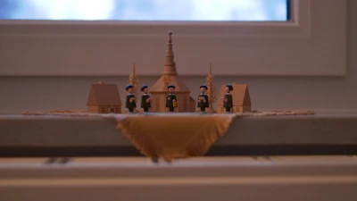 A small, detailed wooden figurine scene featuring miniature houses and people, likely depicting a historical or village setting. The figures are dressed in traditional attire with blue hats and black clothing. The structures include two small houses and one larger building with a steeple, flanked by ornamental sheaves of wheat.