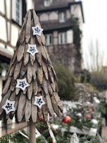 A wooden Christmas tree adorned with star ornaments, set against a blurred background of a traditional European building and holiday decorations. The scene suggests a festive outdoor market or holiday display area.