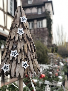 A wooden Christmas tree adorned with star ornaments, set against a blurred background of a traditional European building and holiday decorations. The scene suggests a festive outdoor market or holiday display area.
