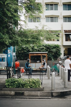 A street scene featuring a few people sitting and conversing in an outdoor area surrounded by greenery. A small street food stall is visible, with individuals interacting and a bicycle parked nearby. The background includes a multi-story building with rectangular windows, and the area is shaded by a large tree.