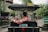 A person wearing a red, floral-patterned shirt is seated in a traditional rickshaw on a city street. The rickshaw is covered with a canopy and has a license plate attached. The background shows a street with greenery and buildings, bustling with people.