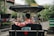 A person wearing a red, floral-patterned shirt is seated in a traditional rickshaw on a city street. The rickshaw is covered with a canopy and has a license plate attached. The background shows a street with greenery and buildings, bustling with people.
