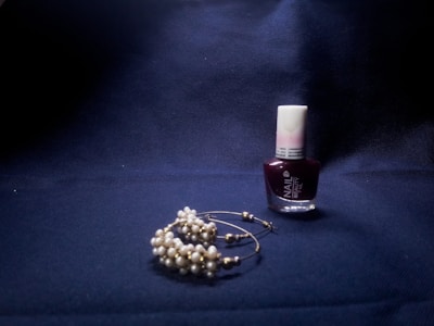 A bottle of deep red nail polish stands upright on a dark, textured fabric surface. Beside it, a delicate pair of earrings adorned with small, light-colored pearls rests on the same surface, casting soft shadows.