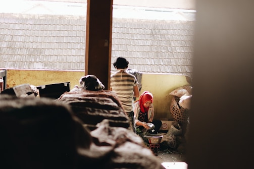 Indoors, two people seem to be involved in their own activities. One is a person in a red headscarf crouching and cleaning vegetables beside a basket. The other individual, seen from the back, stands bending over, appearing to be doing some chore with a piece of cloth. Sunlight filters in through the window above, highlighting the roof outside.