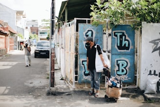 A person wearing a mask and cap is standing beside a cart filled with items, possibly shoes. They are in an urban alleyway next to a wall with graffiti featuring the letters 'Th' and 'B'. Another person in the distance is carrying a package on their shoulder. The scene is lit by daylight.