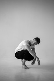 A monochrome image features a man in a plain white t-shirt and dark pants, squatting on a clean, minimalist backdrop. The pose is introspective, with his head bowed and hands resting forward.