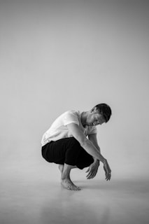 A monochrome image features a man in a plain white t-shirt and dark pants, squatting on a clean, minimalist backdrop. The pose is introspective, with his head bowed and hands resting forward.