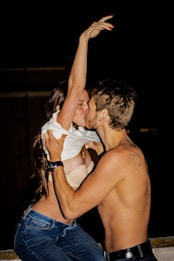 A passionate couple kissing, with the woman raising her arm over her head. The man's shirt is off, revealing a tattoo on his shoulder, while the woman is wearing jeans and a light-colored top that is slightly pulled up.